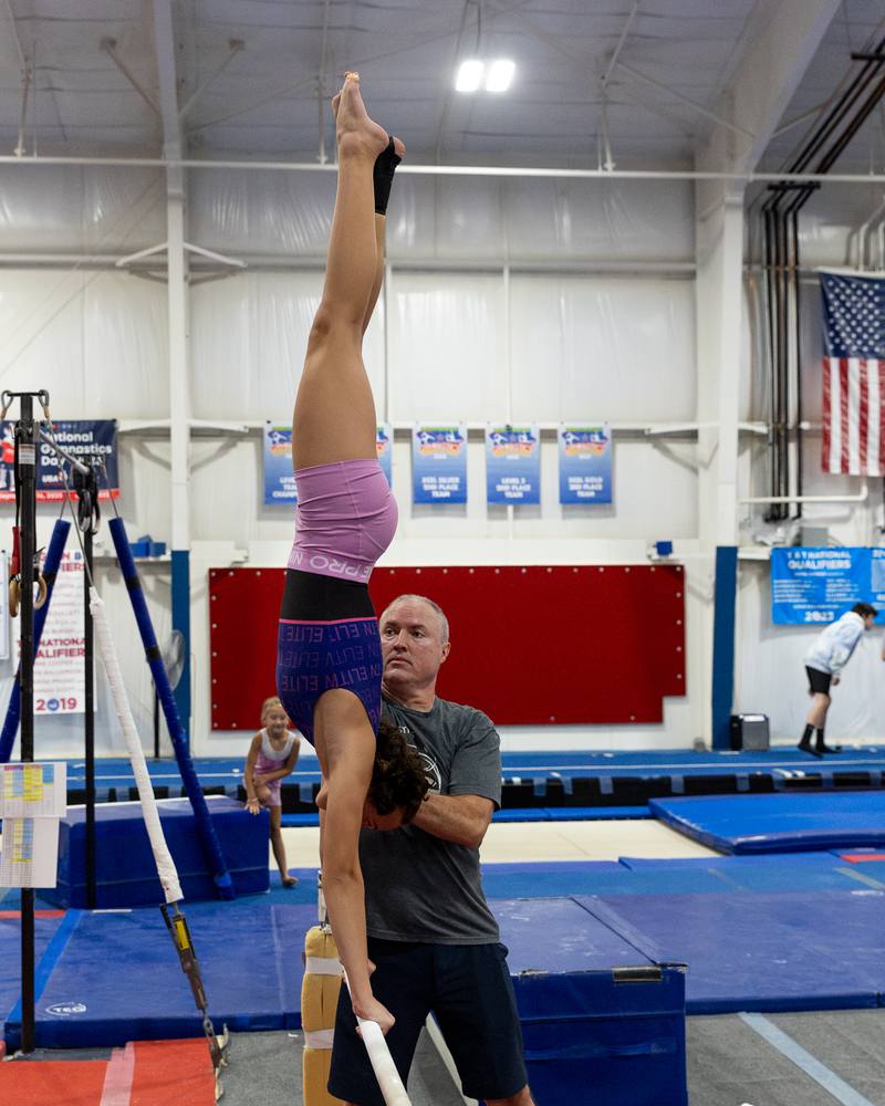 A TEG gymnast holding a handstand while her coach spots, with team banners behind